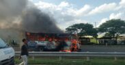 A bus engulfed in flames with thick black smoke rising from it parked on the side of a. A white police vehicle is positioned nearby and a person in casual clothing stands close to the burning bus observing the scene. PHOTO/Kin