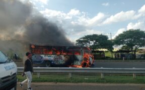 A bus engulfed in flames with thick black smoke rising from it parked on the side of a. A white police vehicle is positioned nearby and a person in casual clothing stands close to the burning bus observing the scene. PHOTO/Kin