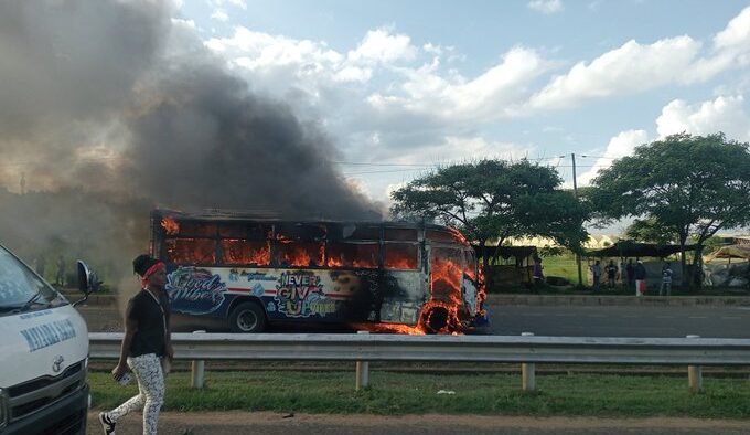 A bus engulfed in flames with thick black smoke rising from it parked on the side of a. A white police vehicle is positioned nearby and a person in casual clothing stands close to the burning bus observing the scene. PHOTO/Kin