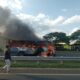 A bus engulfed in flames with thick black smoke rising from it parked on the side of a. A white police vehicle is positioned nearby and a person in casual clothing stands close to the burning bus observing the scene. PHOTO/Kin